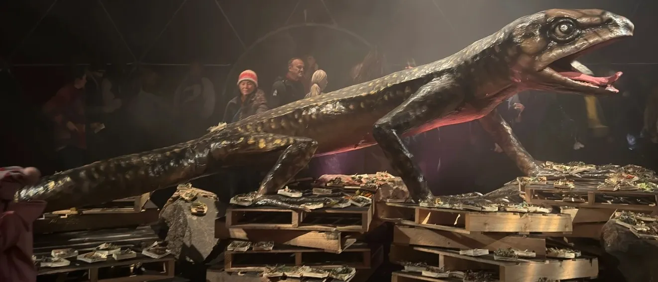 The Ogoh-Ogoh, a giant skink sculpture, crouched on wooden pallets covered in folded paper while visitors look on