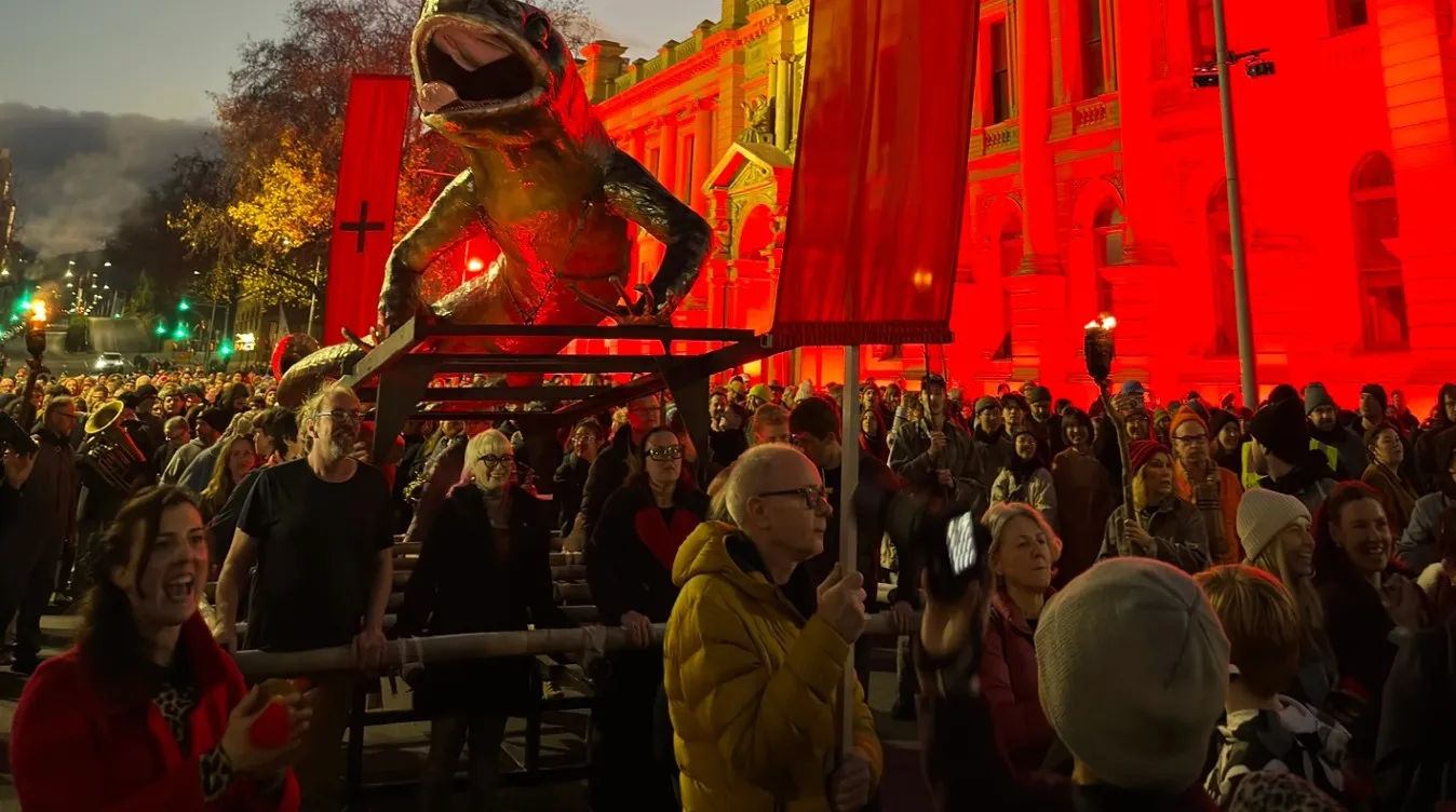 The skink carried through a cheering crowd at dusk, past a sandstone building lit red