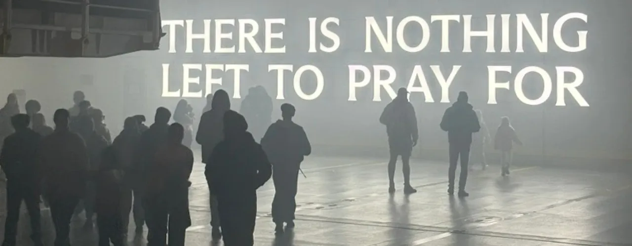 Silhouetted figures walk through a hazy hall beneath glowing words that read There Is Nothing Left To Pray For